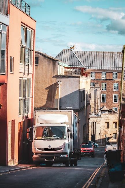 A white delivery van is parked on a narrow street between multi-storey buildings, with the van positioned close to the edge of the pavement. The surrounding buildings feature a mix of brick and plaste